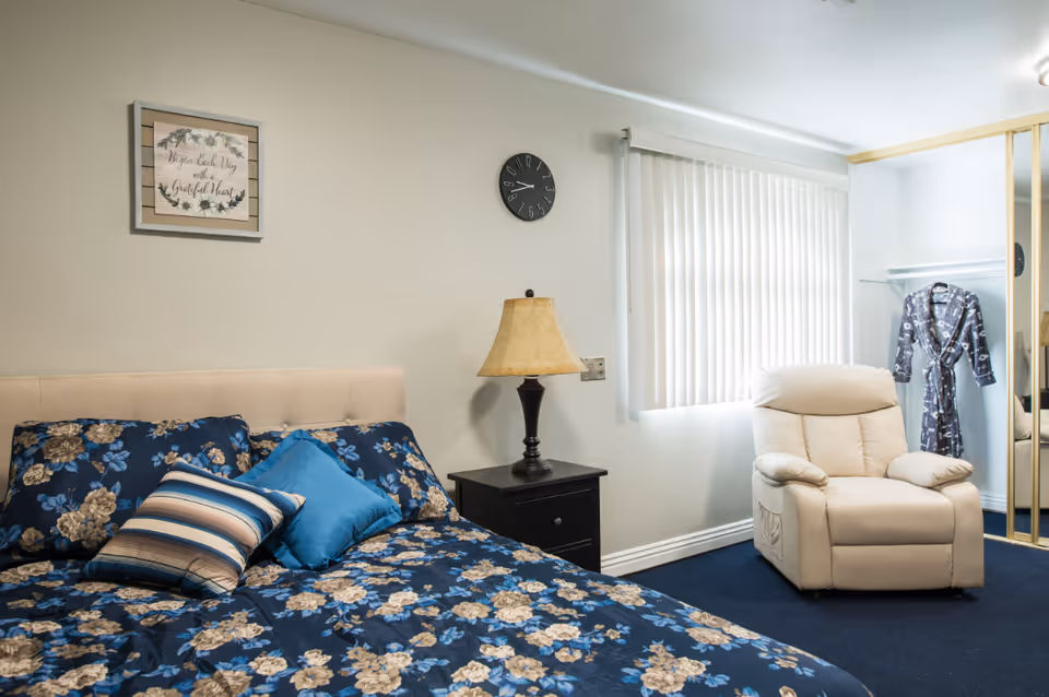 A bedroom with a bed covered in a blue floral bedspread and several pillows. Next to the bed is a black nightstand with a lamp. A beige recliner chair is positioned near a window with vertical blinds. A framed picture hangs on the wall above the bed, and a clock is mounted on the wall above the nightstand. A robe hangs inside an open closet with mirrored sliding doors.