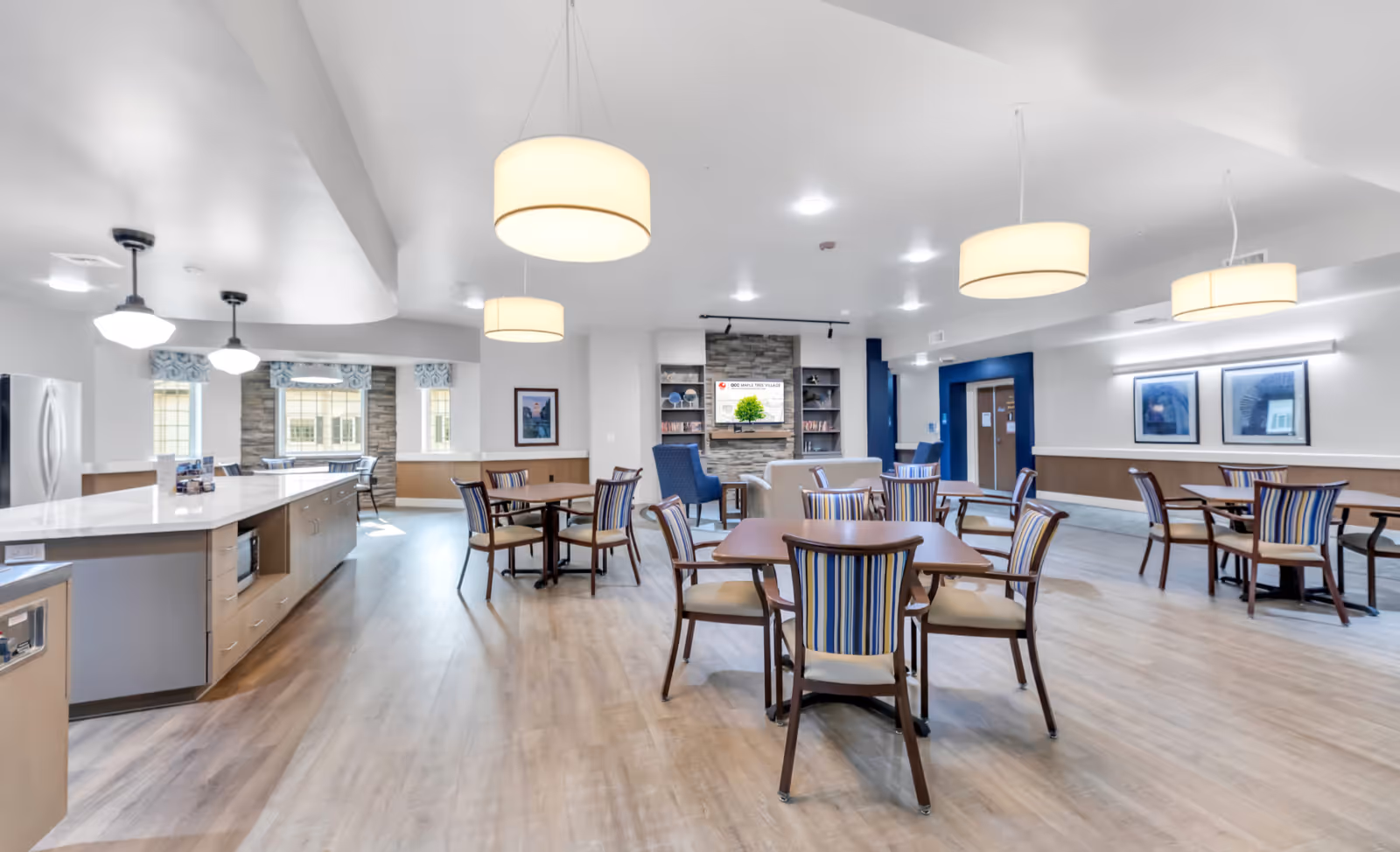 Bright communal dining and lounge area with pendant lights, multiple dining tables and chairs, a kitchen island, and a seating area with a TV.
