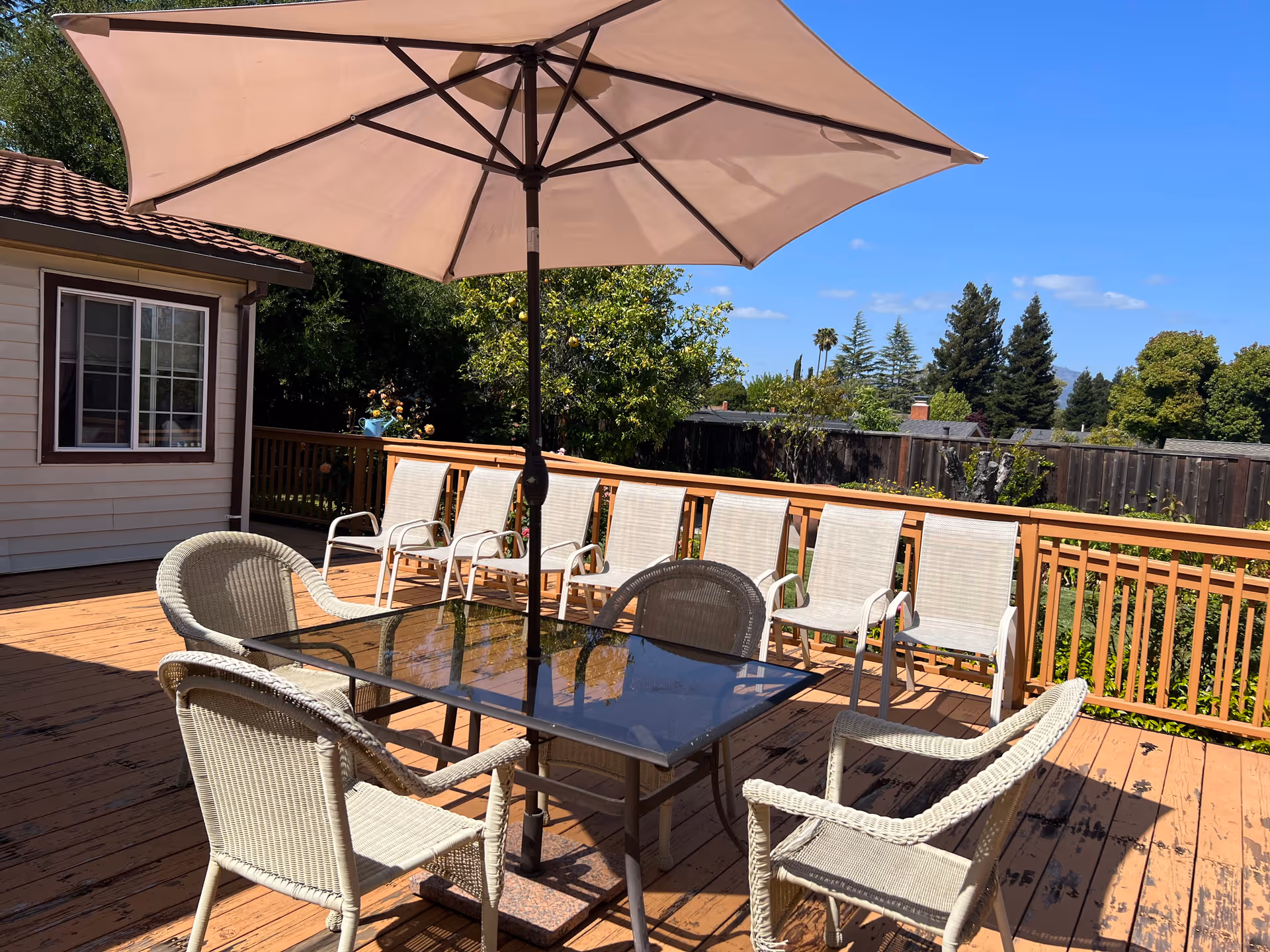 Outdoor wooden deck with a large beige patio umbrella over a glass-top table surrounded by four wicker chairs. Behind the table, there are six white mesh chairs lined up against a wooden railing. Trees and a clear blue sky are visible in the background.