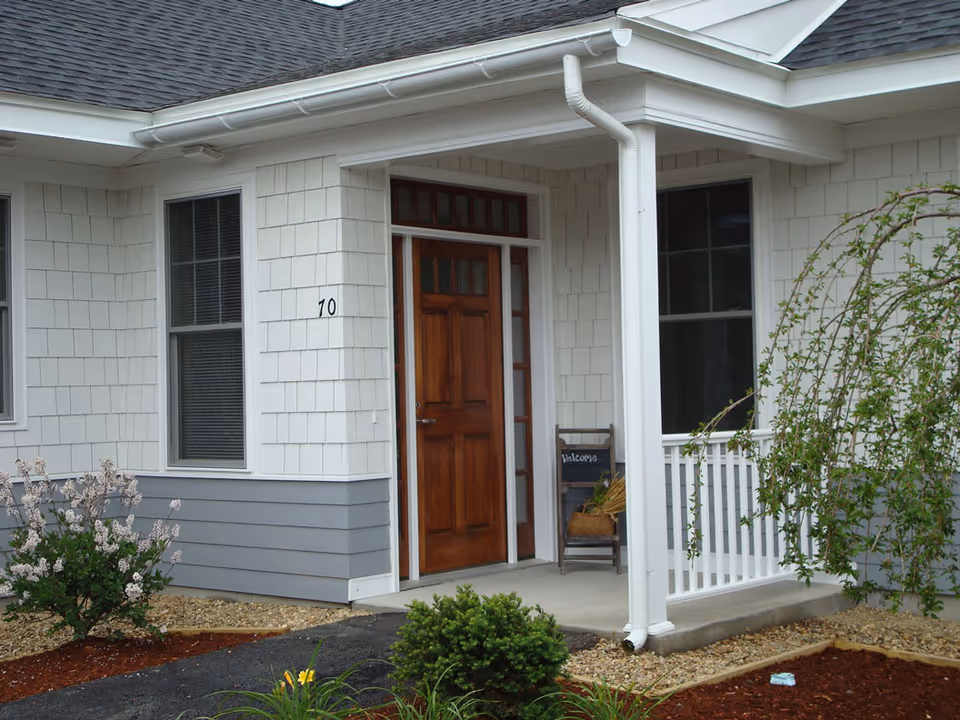Front entrance of a residential building with a wooden door, white siding, two windows, a small porch with a white railing, and a chair with a welcome sign. There are plants and landscaping around the walkway leading to the door.