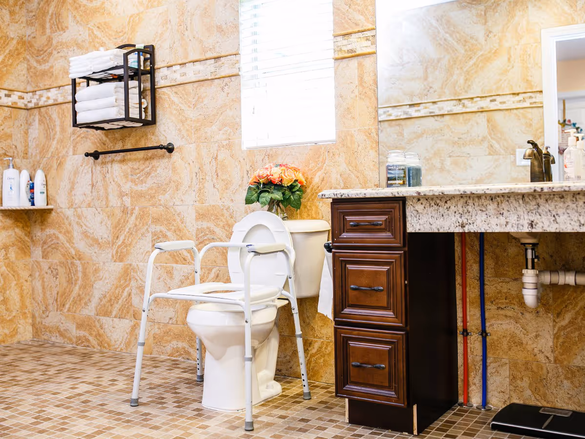 A bathroom with beige marble-patterned walls and tiled floor. There is a white toilet with a white safety frame around it, a wooden cabinet with a granite countertop, a large mirror, and a window with blinds. On the countertop are two candles and a faucet. A shelf with folded white towels and toiletries is mounted on the wall, and a bouquet of flowers is placed on the toilet tank.