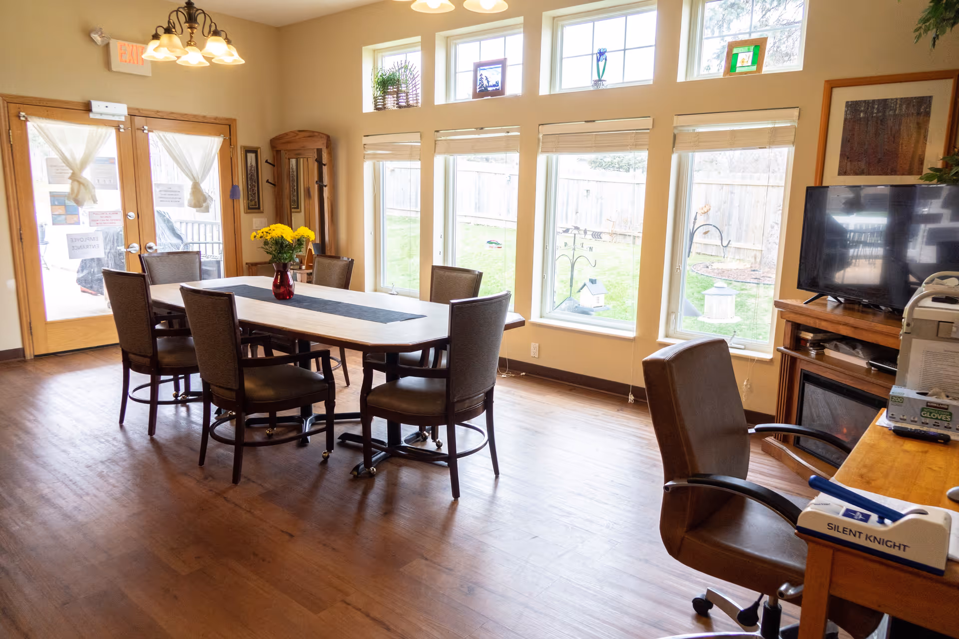 A bright room with large windows overlooking a fenced yard. The room features a wooden table with six chairs around it, a vase with yellow flowers on the table, a TV on a stand, and an office chair near a desk with various items including a box labeled 'Silent Knight'. There are double doors with curtains and an exit sign above them.