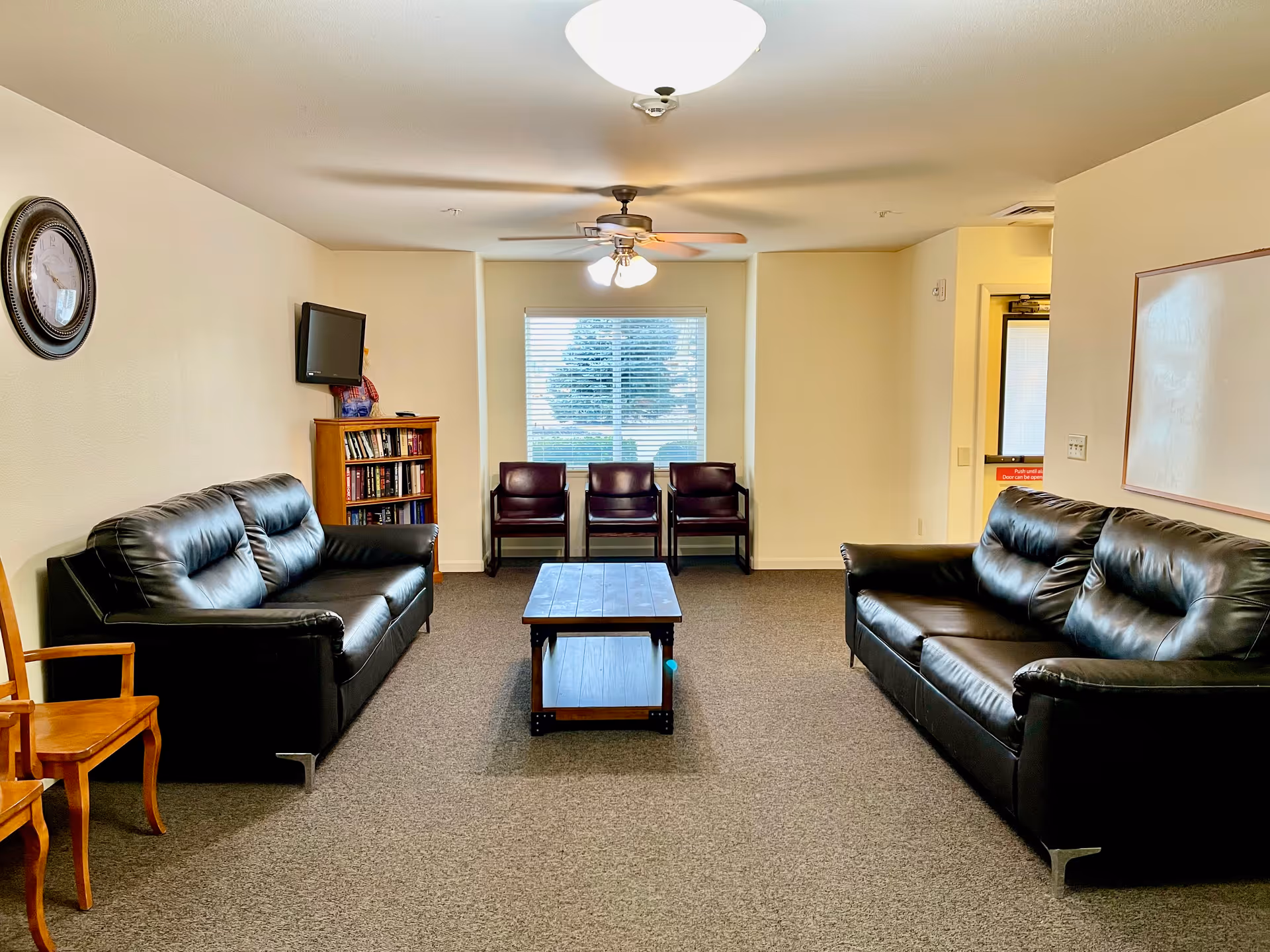 A well-lit living room with two black leather sofas facing each other, a wooden coffee table in the center, three dark chairs against a window with blinds, a wall-mounted TV above a small bookshelf, a wall clock, and a whiteboard on the wall near an exit door.