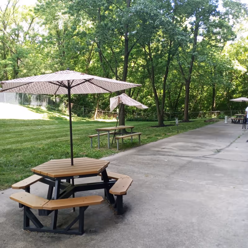 Outdoor area with picnic tables and umbrellas on a concrete path next to a grassy lawn and trees in the background.
