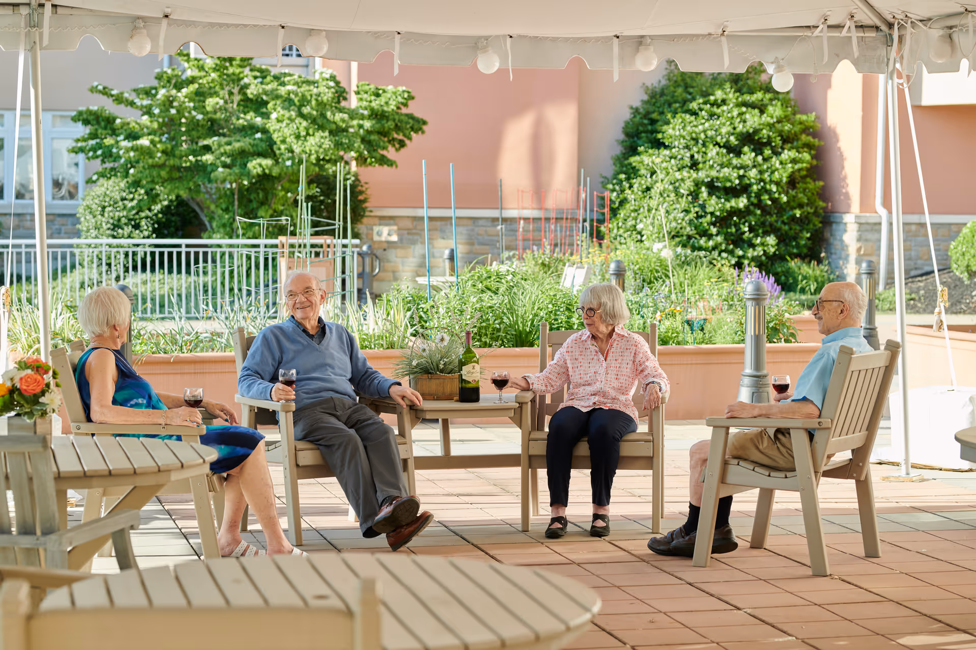 Four elderly people sitting on wooden chairs under a white canopy in an outdoor patio area, enjoying glasses of red wine and engaging in conversation. There are green plants and bushes in the background along with a building wall.