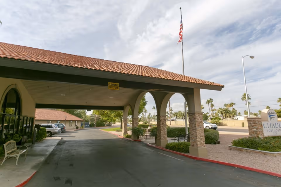 Entrance area of Citadel Post Acute facility with a covered driveway supported by stone pillars, an American flag on a pole, parked cars, and a sign displaying the facility name.
