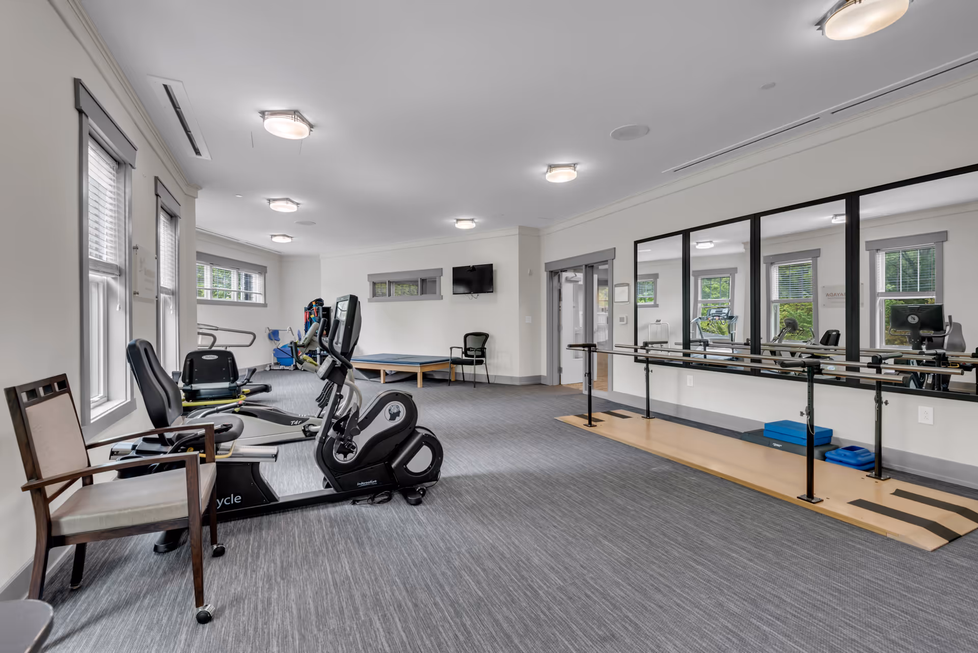 A bright exercise room with large windows, featuring stationary exercise bikes, a ballet barre with mirrors on the wall, a wooden chair, and various exercise equipment. The room has gray carpeting and white walls with ceiling lights.