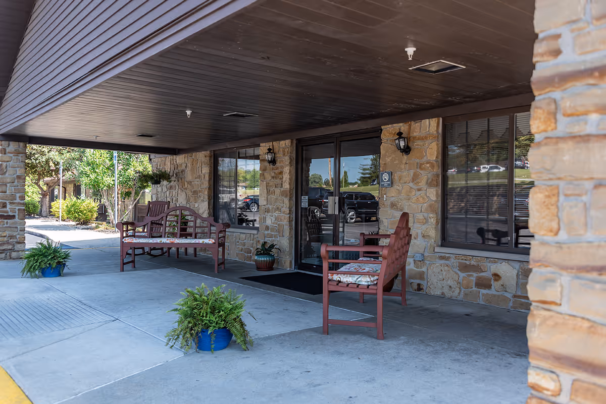 Covered entrance area of Paoli Health & Living Community with stone walls, glass double doors, two red benches with cushions, potted plants, and a view of the parking lot in the background.