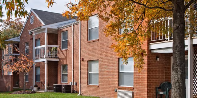 Exterior view of a two-story red-brick building with balconies, a grassy lawn, and trees with autumn foliage.