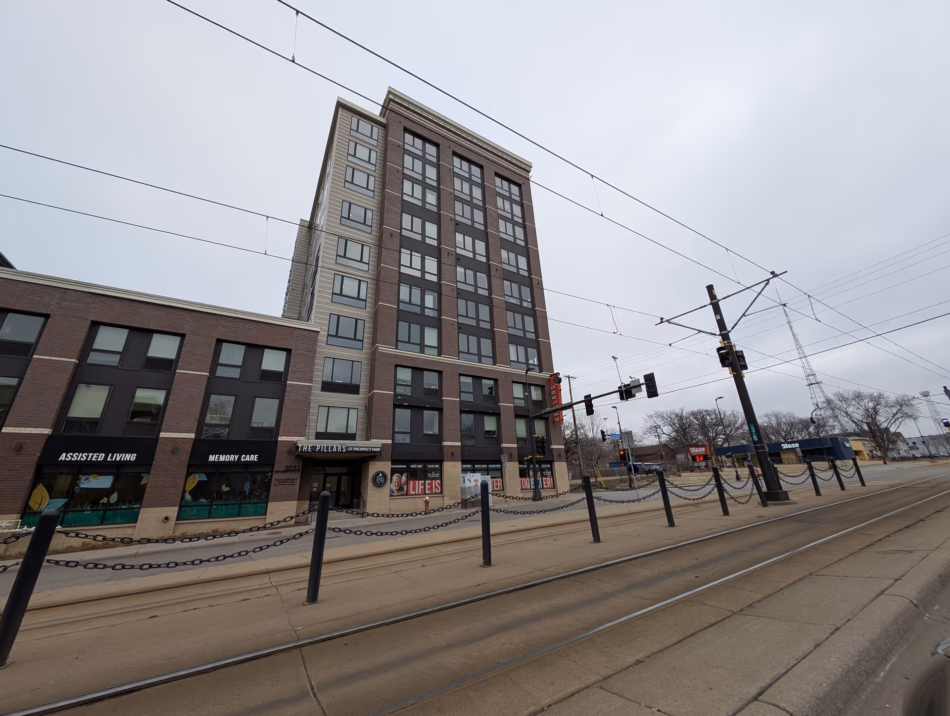 Exterior view of The Pillars of Prospect Park building, a multi-story assisted living and memory care facility with large windows and signage. The street in front has tram tracks, traffic lights, and chain barriers along the sidewalk.