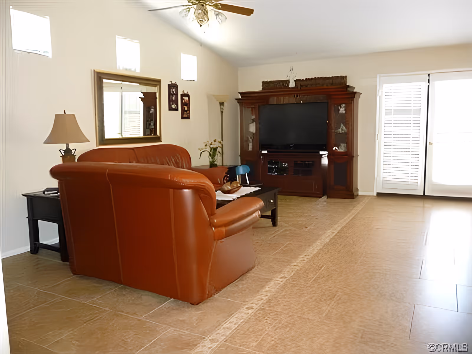 A living room with a brown leather sofa, a wooden coffee table, a large wooden entertainment center with a flat-screen TV, a floor lamp, a table lamp on a side table, a large mirror on the wall, and glass doors with blinds letting in natural light.