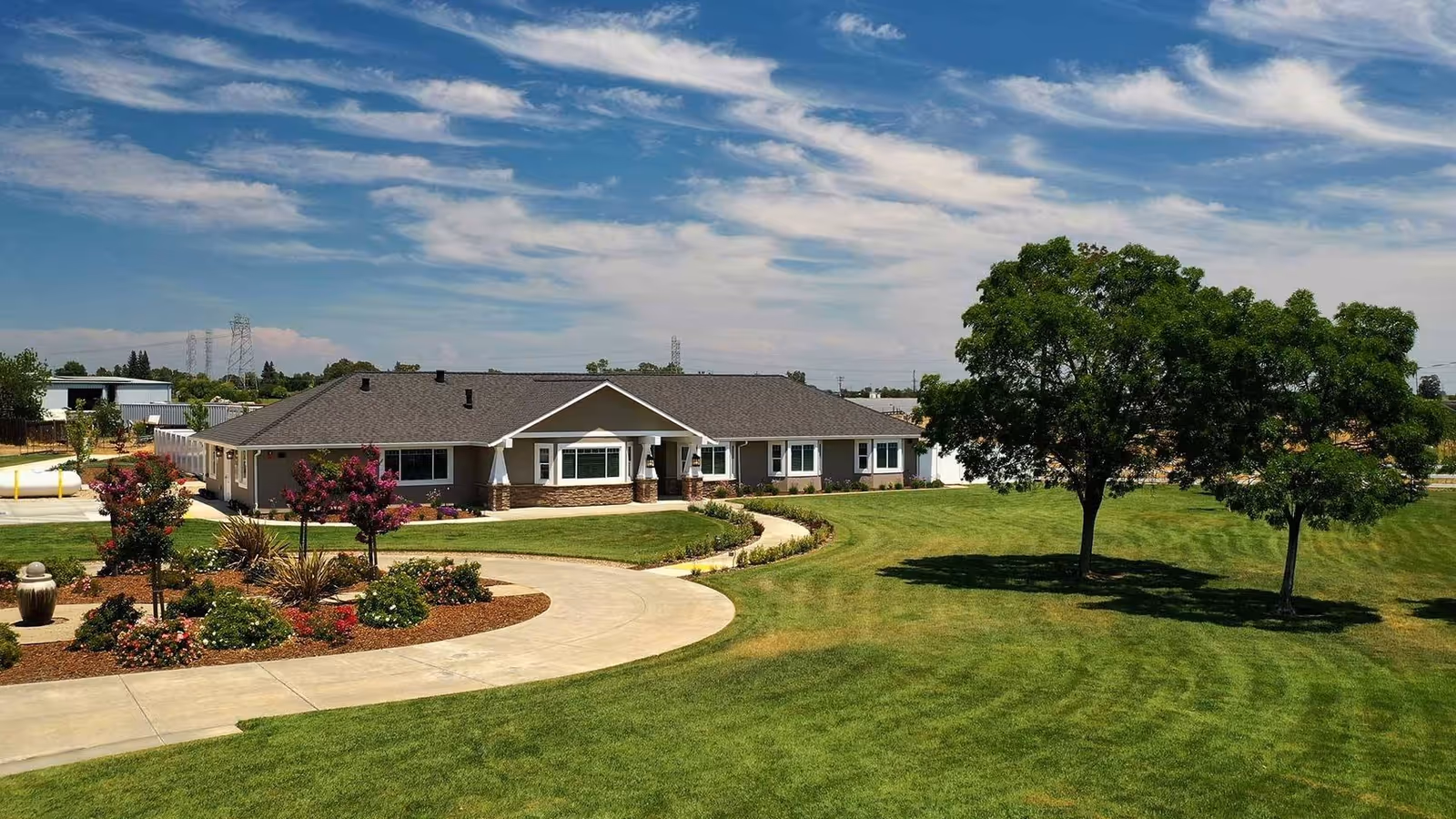 Single-story senior living facility building with a circular driveway, manicured lawn and trees under a partly cloudy sky.