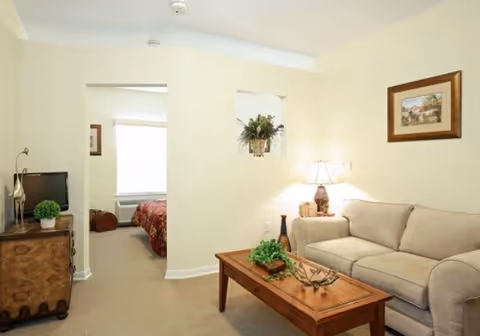 Bright living room with a beige sofa, wooden coffee table, lamp, small TV on a cabinet, and an open doorway leading to a bedroom.
