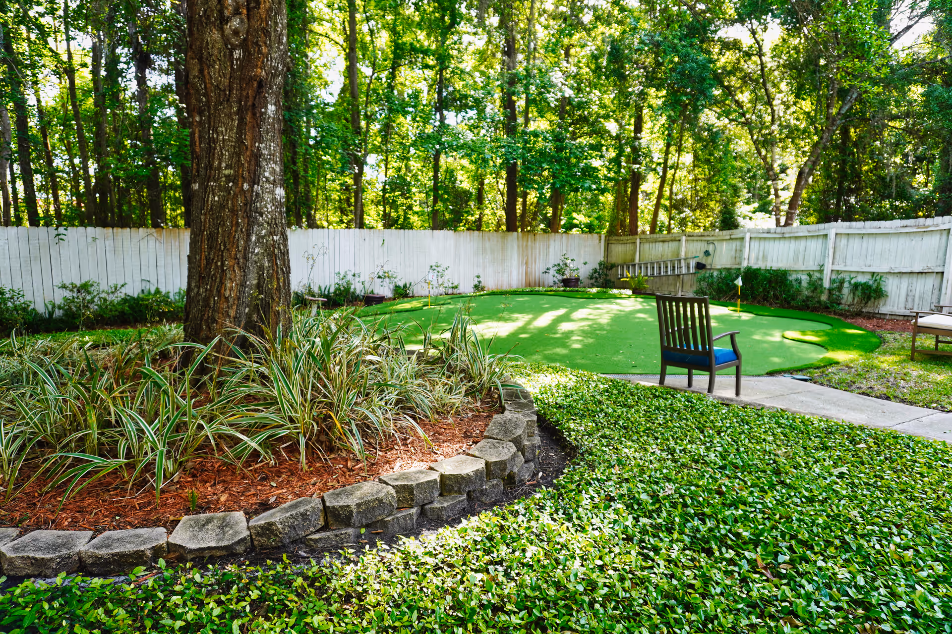 Wooded backyard with a small putting green, a chair, trees, and a white fence.