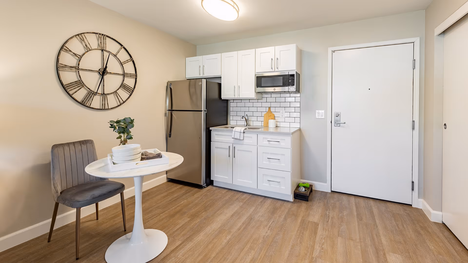 Small kitchen area with white cabinets, a stainless steel refrigerator, microwave, and sink. A round white table with a gray upholstered chair is set against the wall, which has a large decorative clock. The floor is wood, and there is a white door and closet door visible.