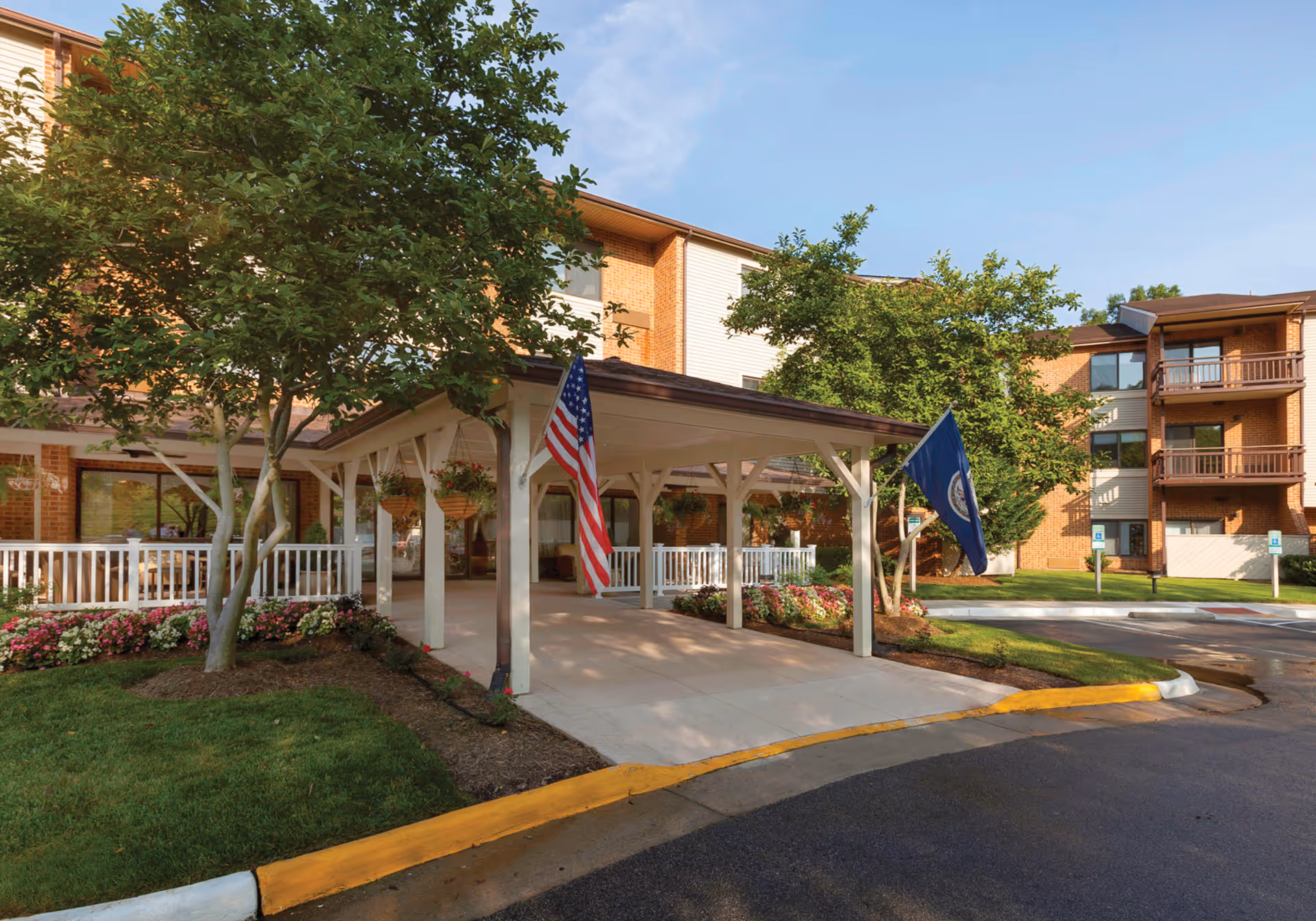 Entrance area of Potomac Place Assisted Living and Memory Care facility with a covered drop-off area, two flags (American and another flag), landscaped garden beds with flowers, trees, and a multi-story brick building in the background under a clear blue sky.