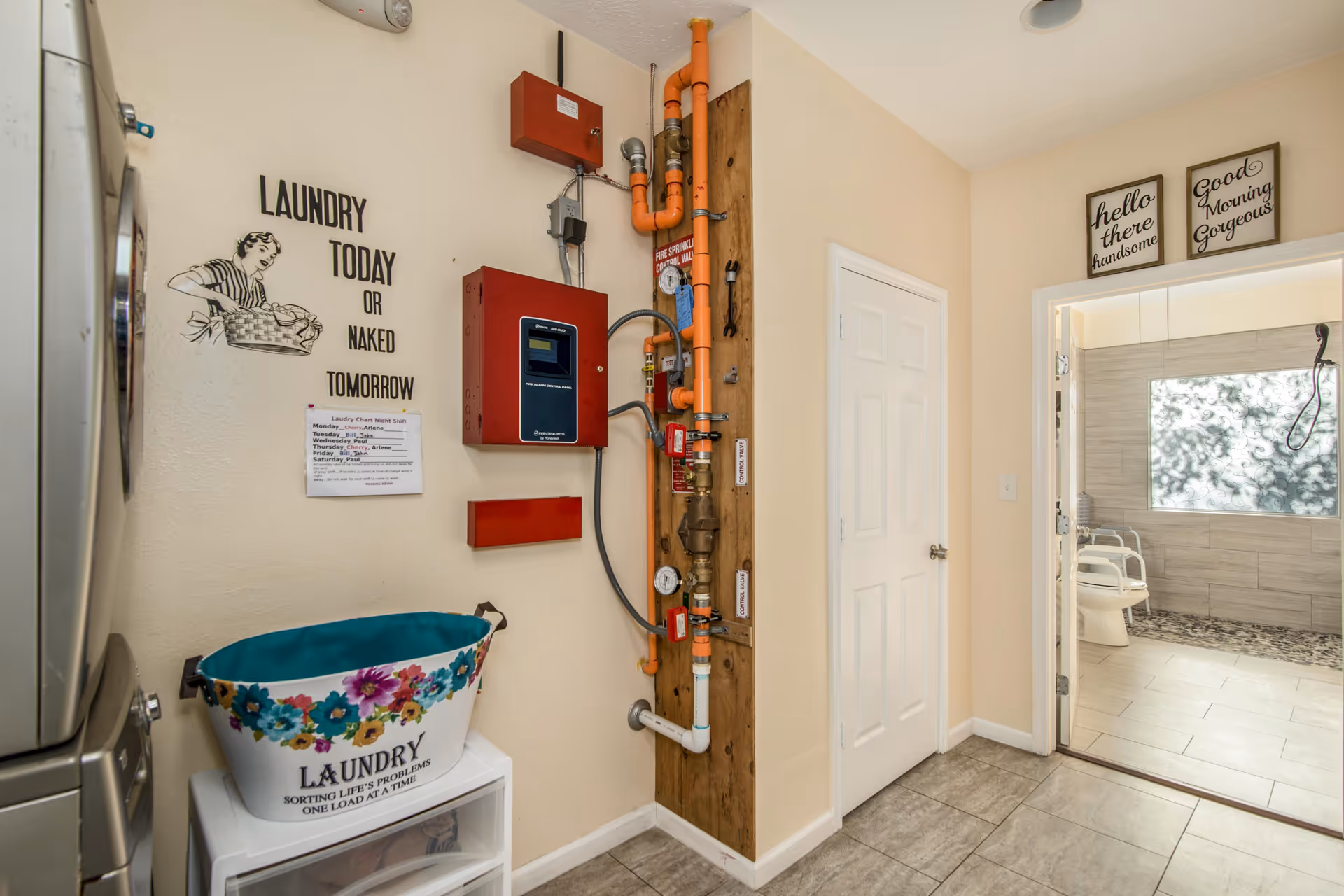 Laundry room with a washing machine on the left, a floral laundry basket on a white storage unit, and a wall with plumbing pipes and fire sprinkler system components. A humorous sign on the wall reads 'Laundry today or naked tomorrow.' Through an open door, a bathroom with a walk-in shower and a chair is visible. Two framed signs above the bathroom door say 'hello there handsome' and 'Good Morning Gorgeous.'