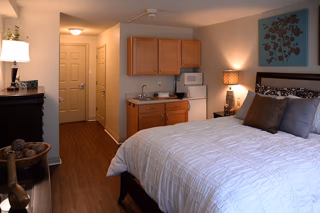 A cozy bedroom in a senior living facility featuring a large bed with white bedding and multiple pillows. To the right of the bed is a nightstand with a lamp and decorative items. Above the bed hangs a blue artwork with floral design. On the left side of the room, there is a small kitchenette with wooden cabinets, a sink, a microwave, and a mini refrigerator. The floor is wooden, and the walls are painted light beige. A hallway with closed doors is visible in the background.