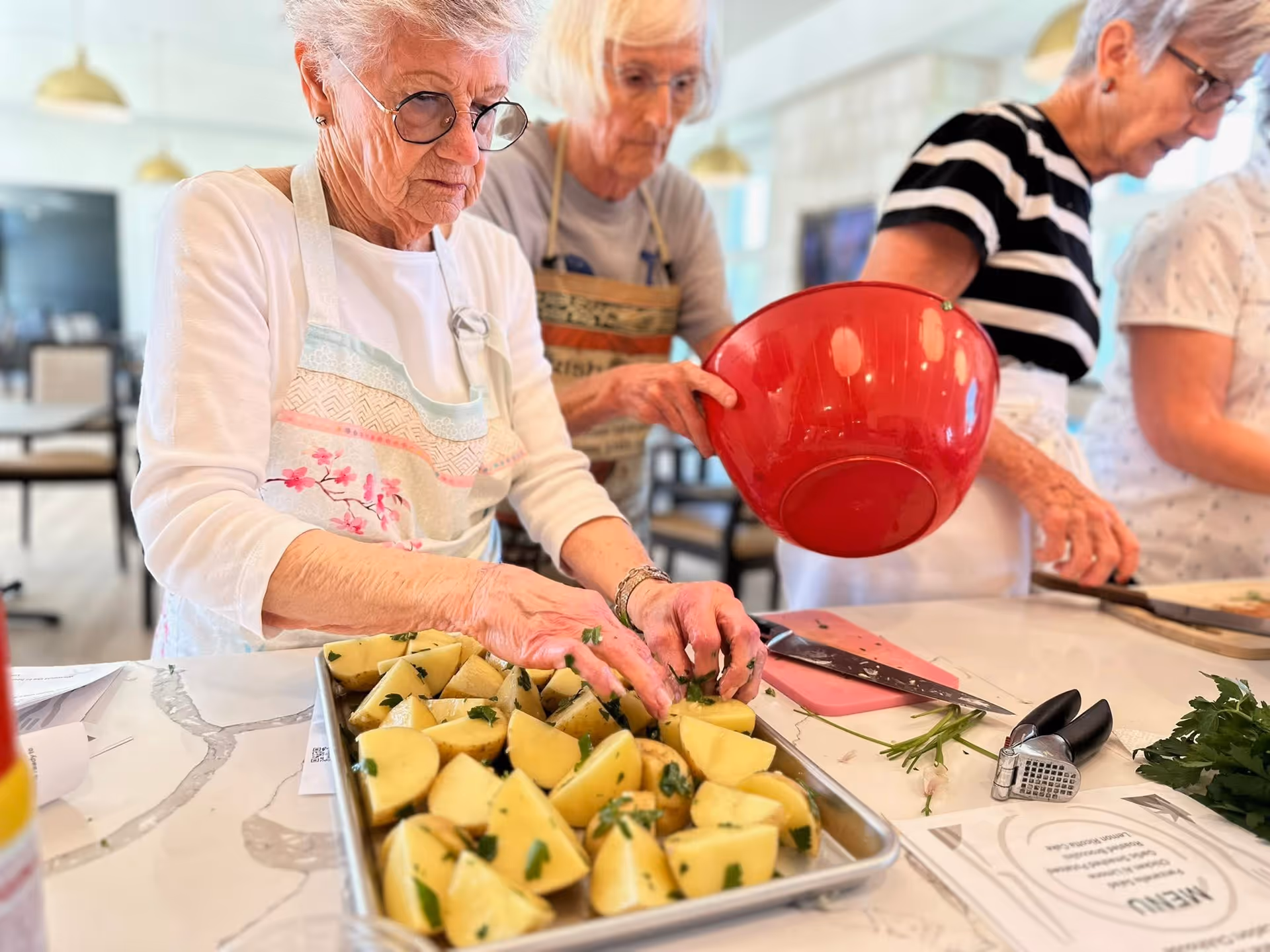 Three elderly women preparing food together in a kitchen, one arranging cut potatoes on a baking tray while another holds a large red mixing bowl, and the third working on a cutting board. The kitchen counter has various utensils and fresh herbs.