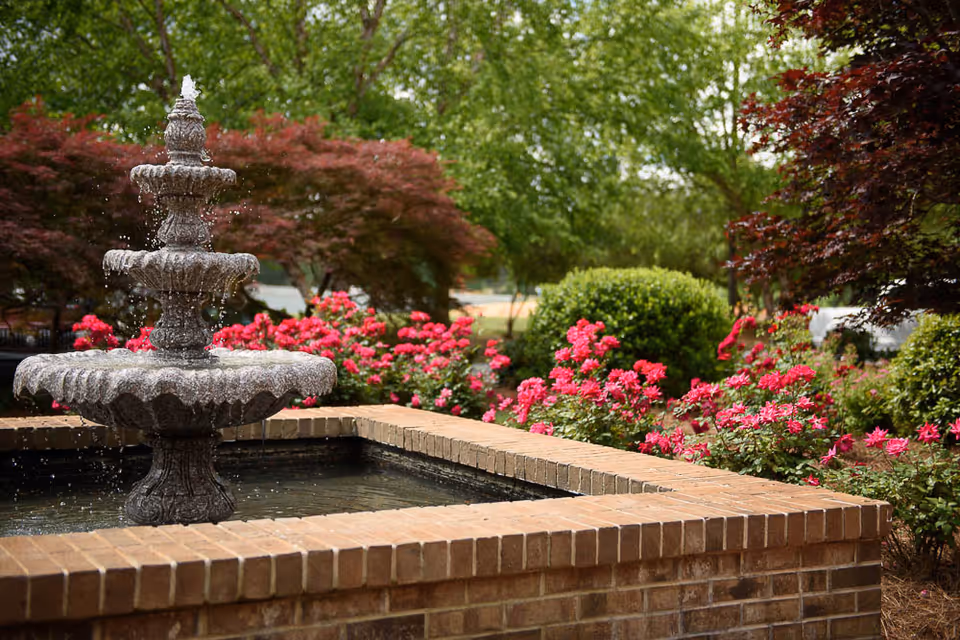 A tiered stone water fountain surrounded by a brick border, with vibrant pink flowers and green bushes in the background under a canopy of trees.
