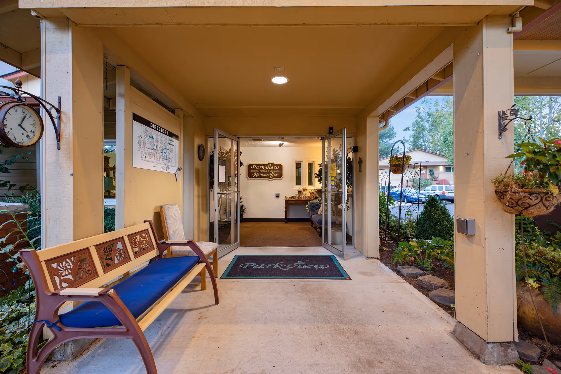 Covered entrance area of Parkview Christian Retirement Community with a wooden bench with blue cushion on the left, glass double doors leading inside, a directory on the wall, and plants hanging and growing around the entrance.