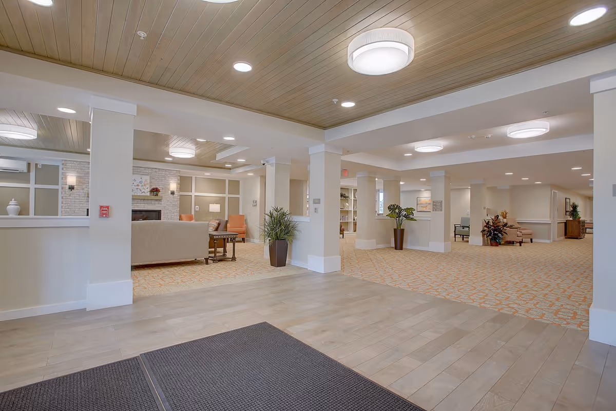 Spacious and well-lit interior common area with beige walls, wooden ceiling, recessed lighting, and a mix of tiled and carpeted flooring. The space includes seating areas with sofas and chairs, potted plants, and a fireplace in the background.