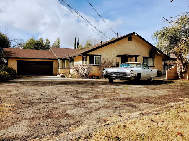 Single-story beige stucco house with a large paved driveway and a vintage white car parked in front under a partly cloudy sky.