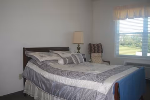 A simple furnished bedroom with a made double bed, bedside lamp, upholstered chair, and a window with blinds showing greenery outside.