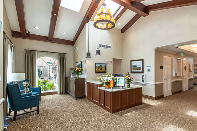 A bright and welcoming interior space with high ceilings and exposed wooden beams. There is a chandelier and pendant lights hanging from the ceiling. The room features a reception desk with flowers and framed pictures on it. To the left, there is a large window with curtains and a blue armchair next to a small table with a lamp and flowers. The carpet has a patterned design, and the walls are painted in neutral tones with some artwork hanging on them. A hallway extends to the right with doors and additional lighting.