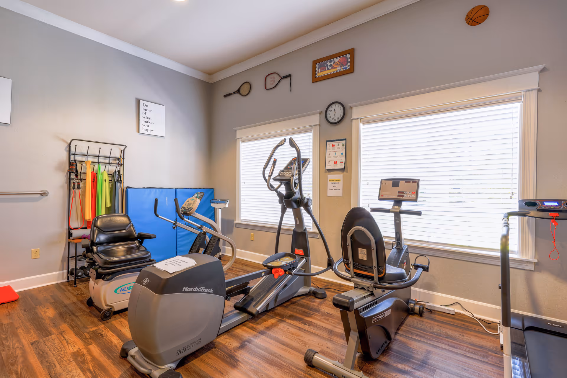A small fitness room with exercise equipment including a recumbent bike, elliptical machine, rowing machine, and treadmill. The room has wooden flooring, two large windows with blinds, and light gray walls decorated with framed pictures and two tennis rackets mounted above the windows. There is also a rack with resistance bands and other exercise accessories.