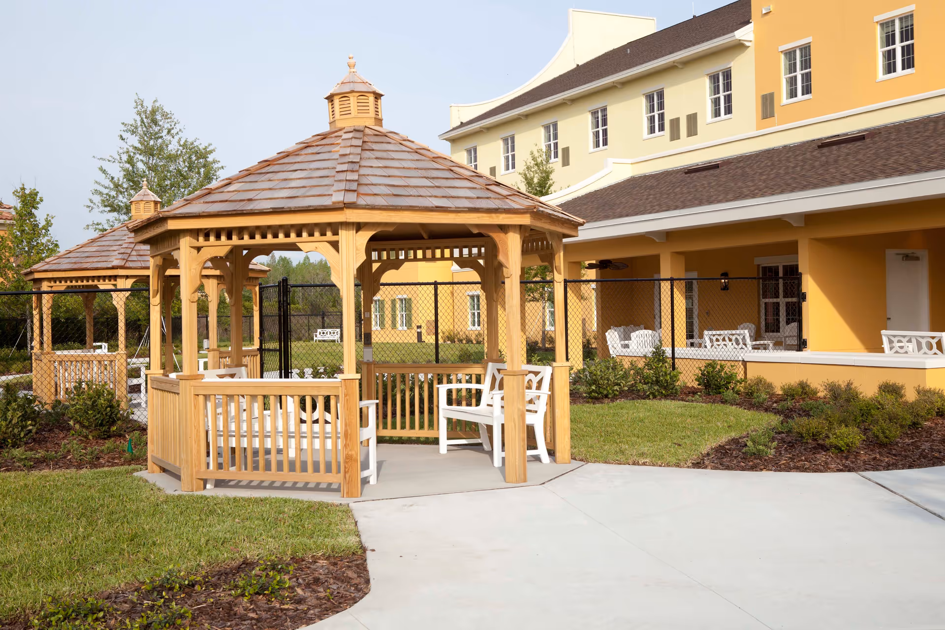 Wooden gazebo with white chairs on a paved patio in front of a yellow senior living building.