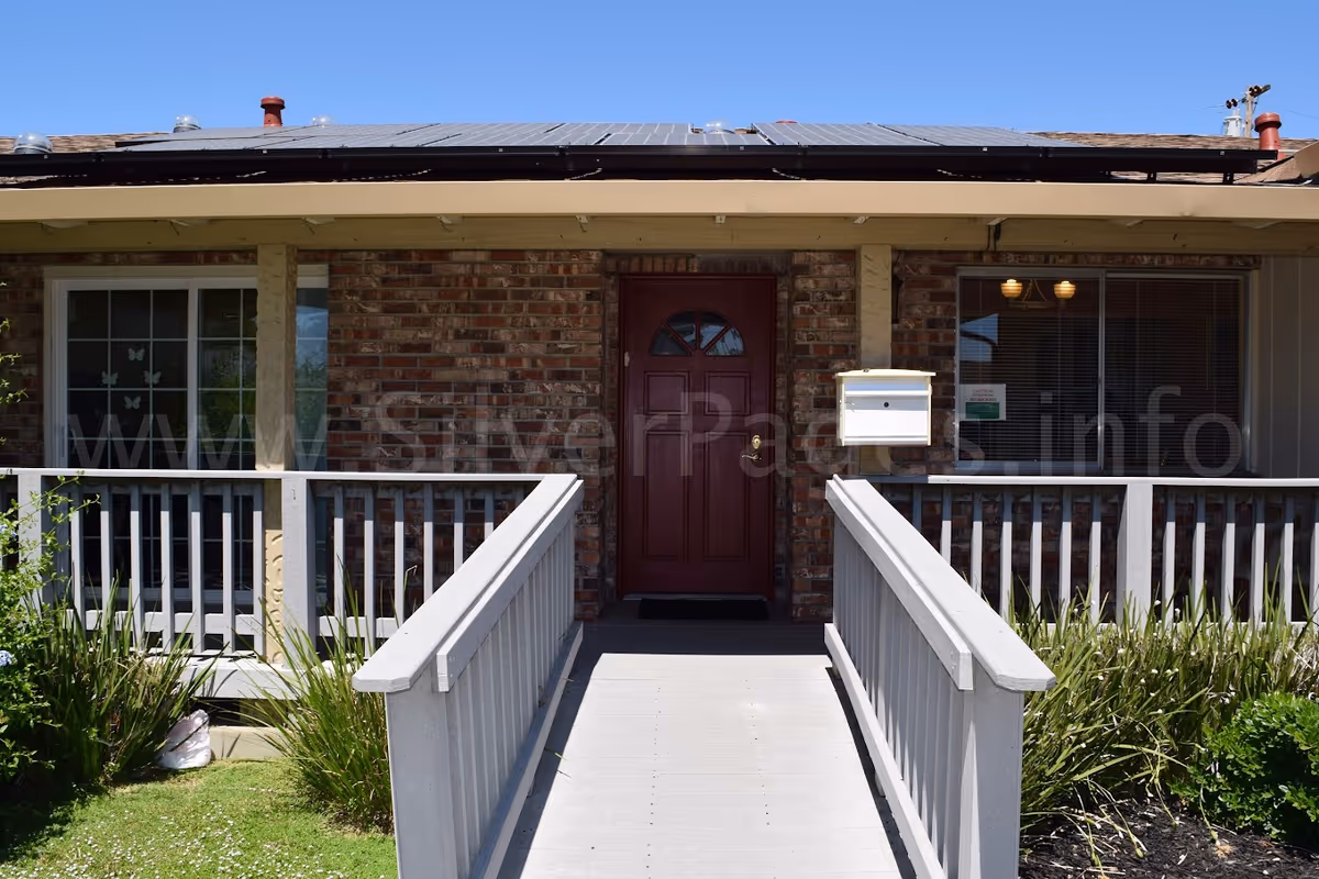 Front entrance of a single-story brick building with a wheelchair ramp leading to a red door, porch railings, mailbox, and windows.