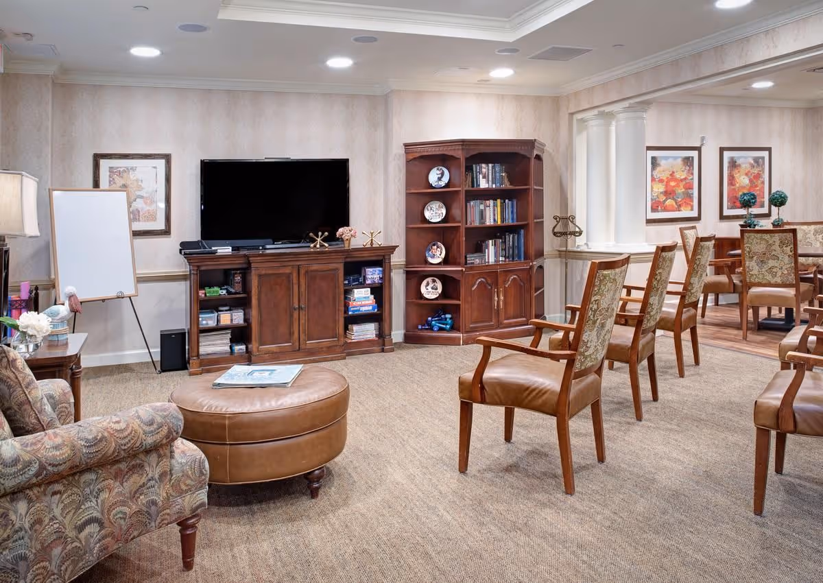 A warmly furnished common living room with a TV on a wooden console, bookshelves, an ottoman and several wooden chairs arranged for seating.