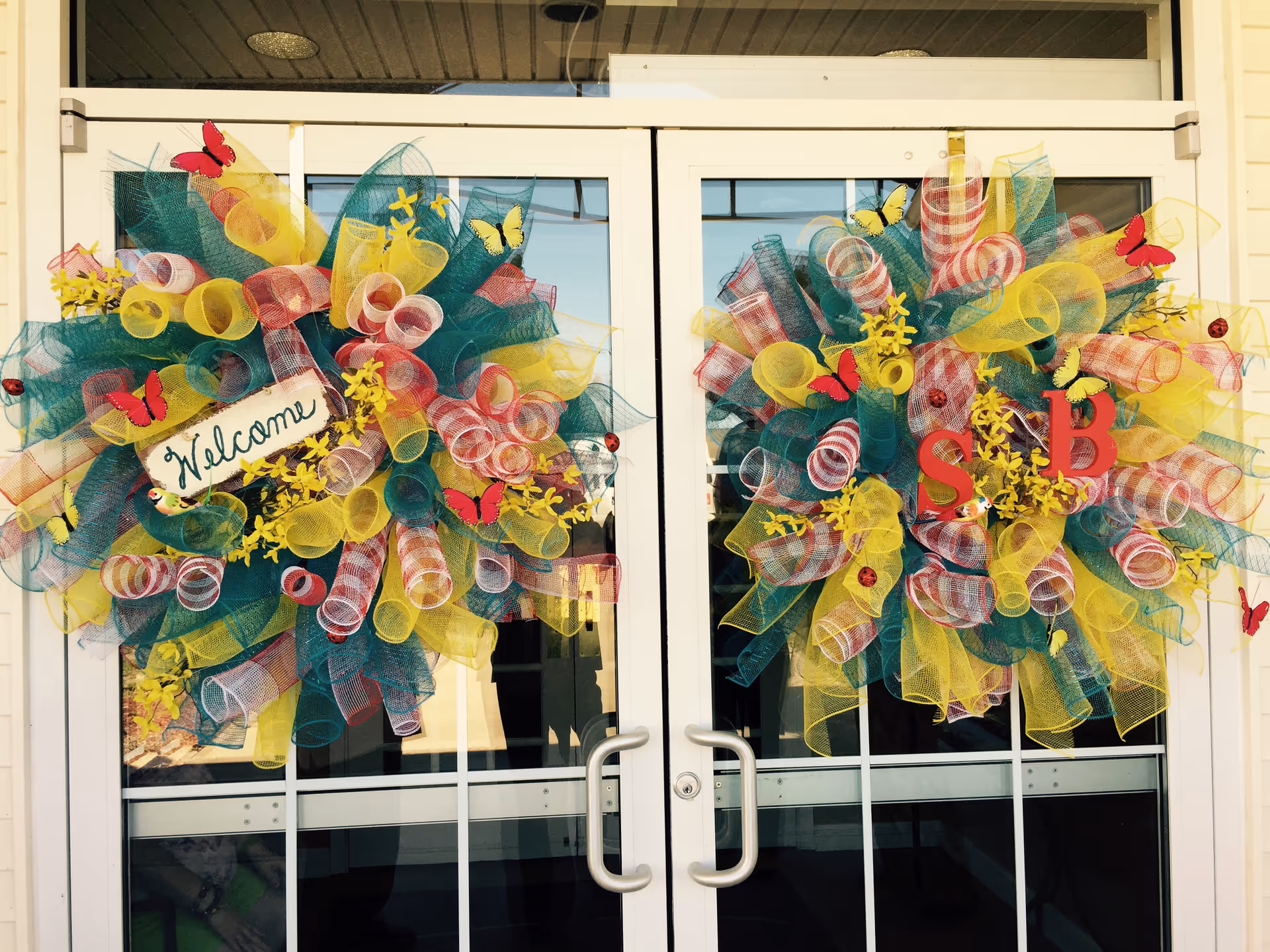 Double glass doors decorated with two large colorful wreaths made of yellow, green, and red mesh ribbons, adorned with red and yellow butterflies and small yellow flowers. One wreath has a sign that says 'Welcome' and the other has red letters 'S' and 'B'.