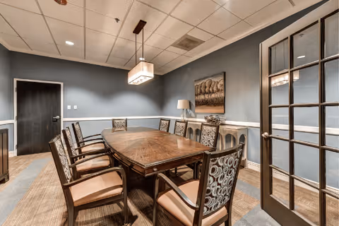 A formal dining room with a large wooden table surrounded by eight cushioned chairs. The walls are painted blue with white trim, and there is a modern rectangular ceiling light fixture above the table. A sideboard with a lamp and decorative bowl is against the far wall, which also features a framed landscape painting. The room has a carpeted floor and a glass-paneled door on the right side.