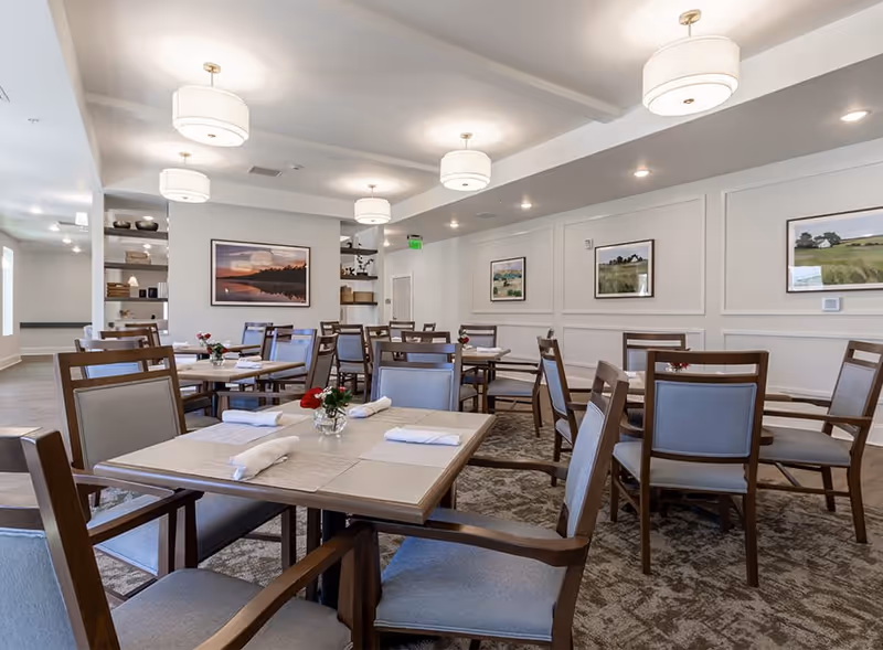 A bright and spacious dining room with multiple wooden tables and chairs arranged neatly. Each table is set with placemats, napkins, and small vases containing red flowers. The walls are decorated with framed landscape paintings, and the ceiling has several modern light fixtures. The room has a clean and welcoming atmosphere.