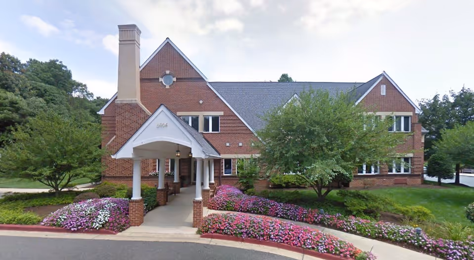 Front exterior view of a two-story brick building with a covered entrance supported by white columns. The building is surrounded by well-maintained landscaping including colorful flower beds and green trees.