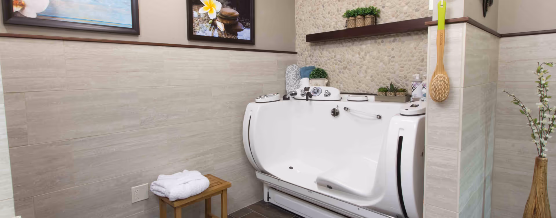 A modern walk-in bathtub in a bathroom with beige tiled walls and a pebble-textured accent wall. The bathtub has built-in controls and a seat. There is a wooden stool with folded white towels next to the tub, a wooden bath brush hanging on the wall, and decorative plants on a shelf and in a vase. Two framed pictures with floral and water themes hang above the tub.