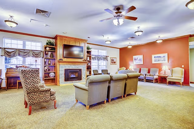 A spacious living room area with beige carpet and multiple seating options including three armchairs facing a fireplace with a mounted flat-screen TV above it. There are built-in wooden shelves on either side of the fireplace with decorative items. The room has large windows with valance curtains, ceiling fans with lights, and several framed pictures on the walls. Additional chairs are arranged along the far wall.