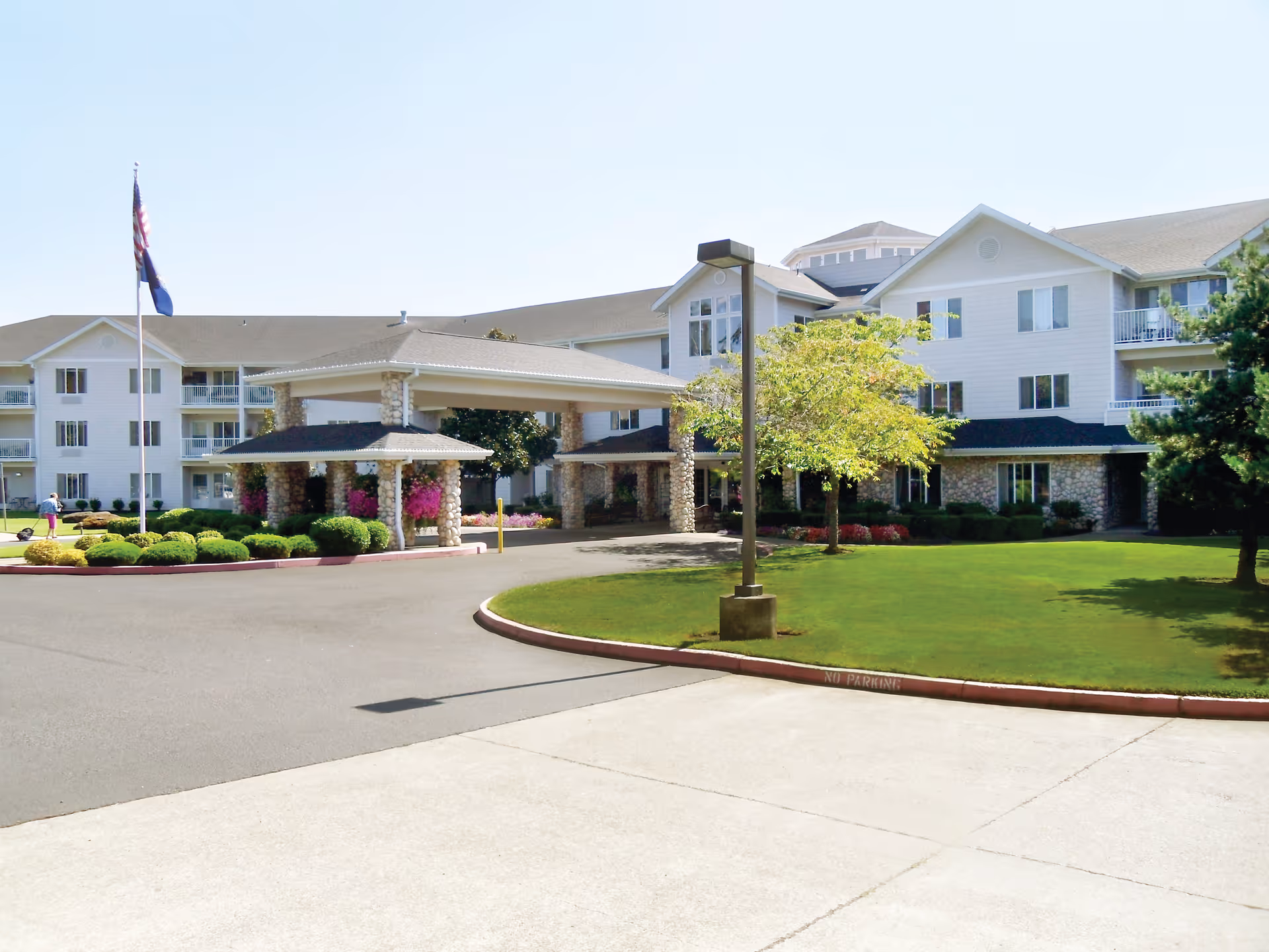 Front entrance of a multi-story senior living building with a porte-cochere, flagpoles, and landscaped lawn.