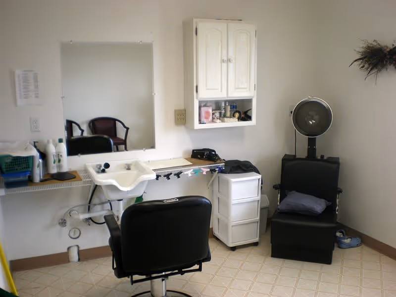 Interior of a small hair salon or barber area with a black salon chair facing a sink and mirror. There is a white cabinet mounted on the wall, a small white drawer unit, and a black hair dryer chair with a hood dryer. A pair of slippers is on the floor near the corner.