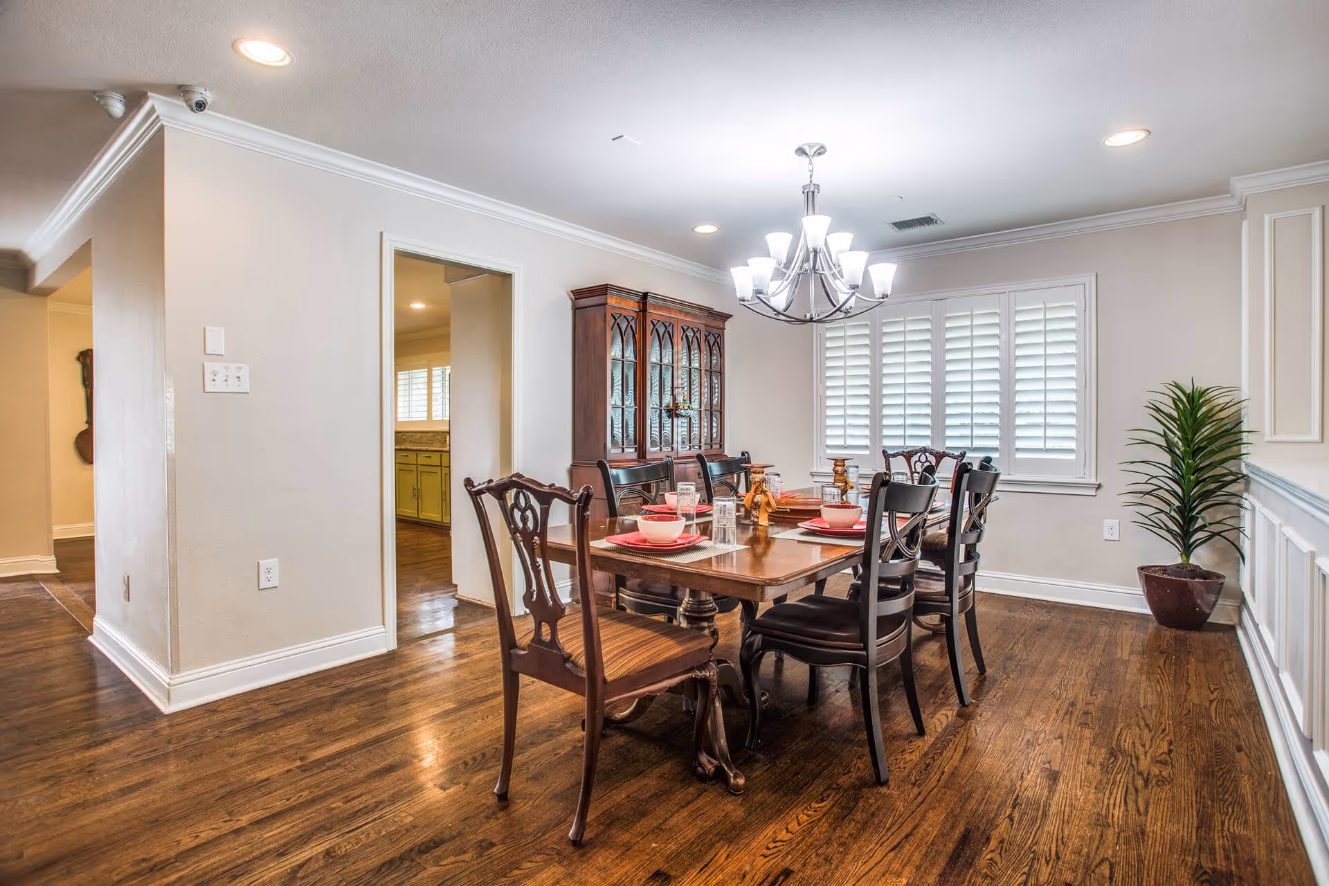 A well-lit dining room with a wooden dining table set for six, featuring a mix of dark wooden chairs. A chandelier with multiple lights hangs above the table. There is a wooden china cabinet against the wall and a potted plant in the corner. The room has hardwood floors and white walls with crown molding and wainscoting. A window with white plantation shutters is visible on the far wall.