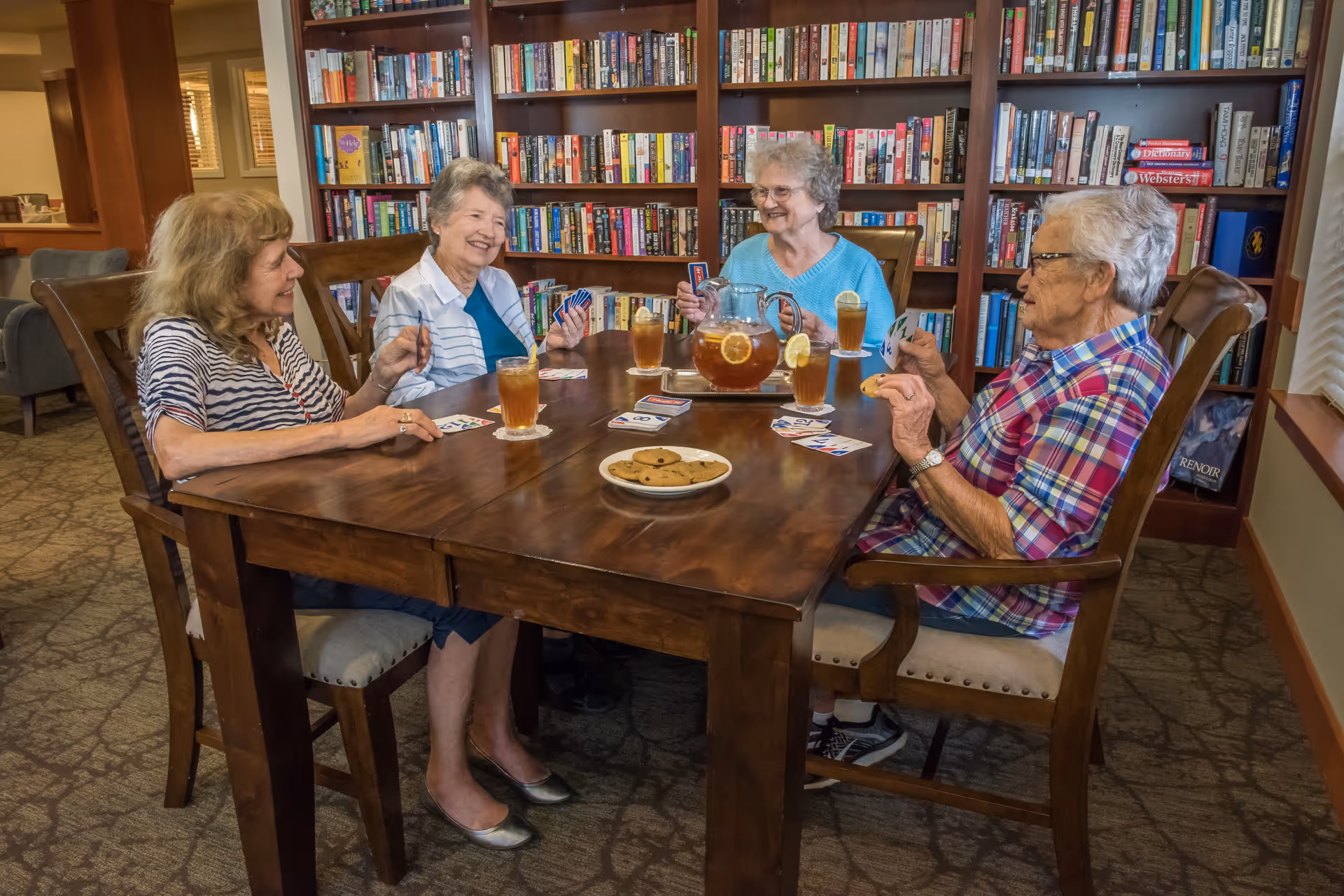 Four elderly women sitting around a wooden table playing cards in a room with a large bookshelf filled with books. On the table are glasses of iced tea, a pitcher of iced tea with lemon slices, and a plate of cookies. The room has carpeted floors and warm lighting.
