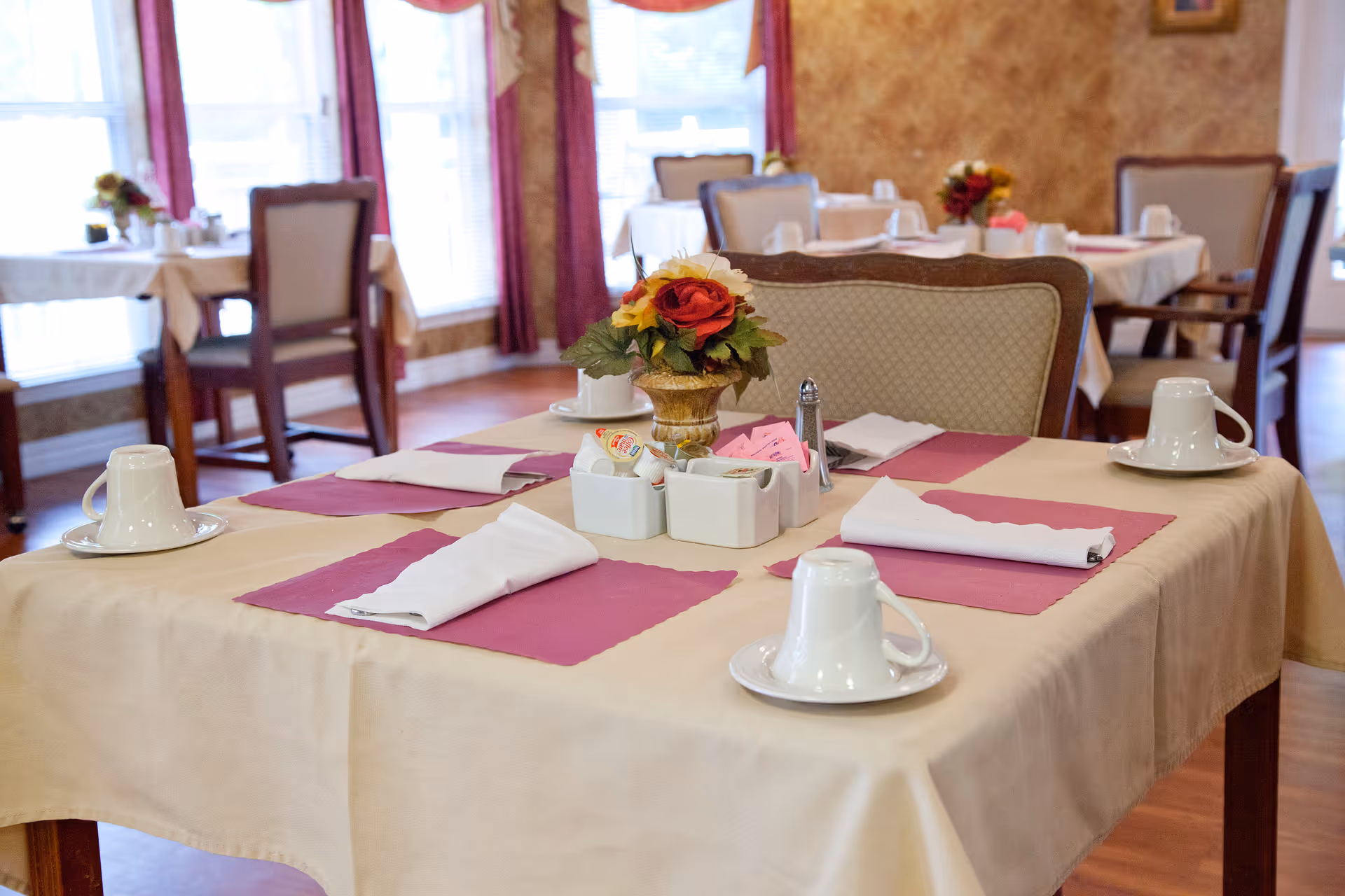 A dining table set for four with beige tablecloth and purple placemats, white folded napkins, white cups on saucers, and a small centerpiece with artificial flowers in a vase. The background shows more tables and chairs in a warmly lit dining room with large windows and curtains.