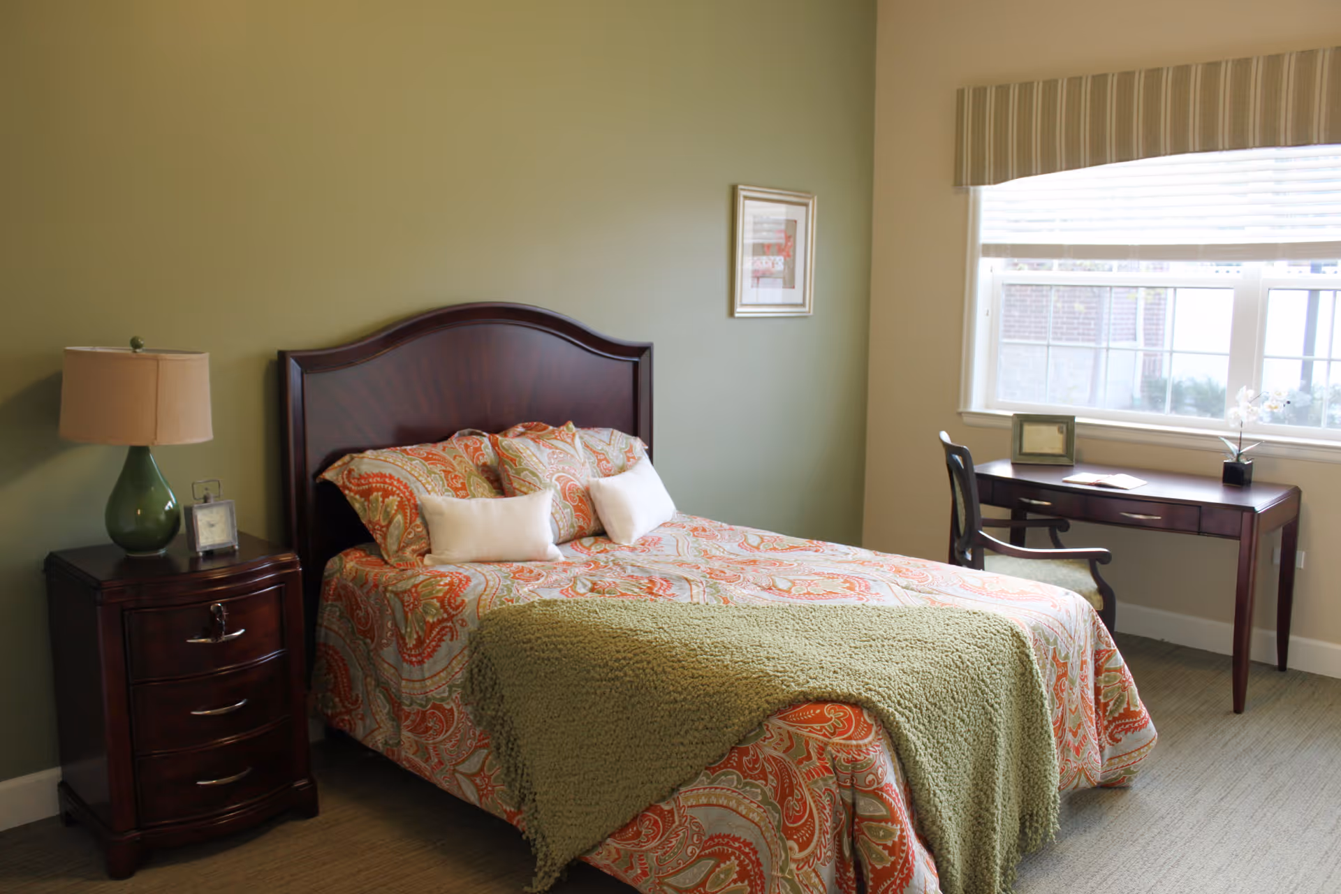 Bedroom with a patterned bed and wooden headboard, a nightstand with lamp, and a desk by a window.