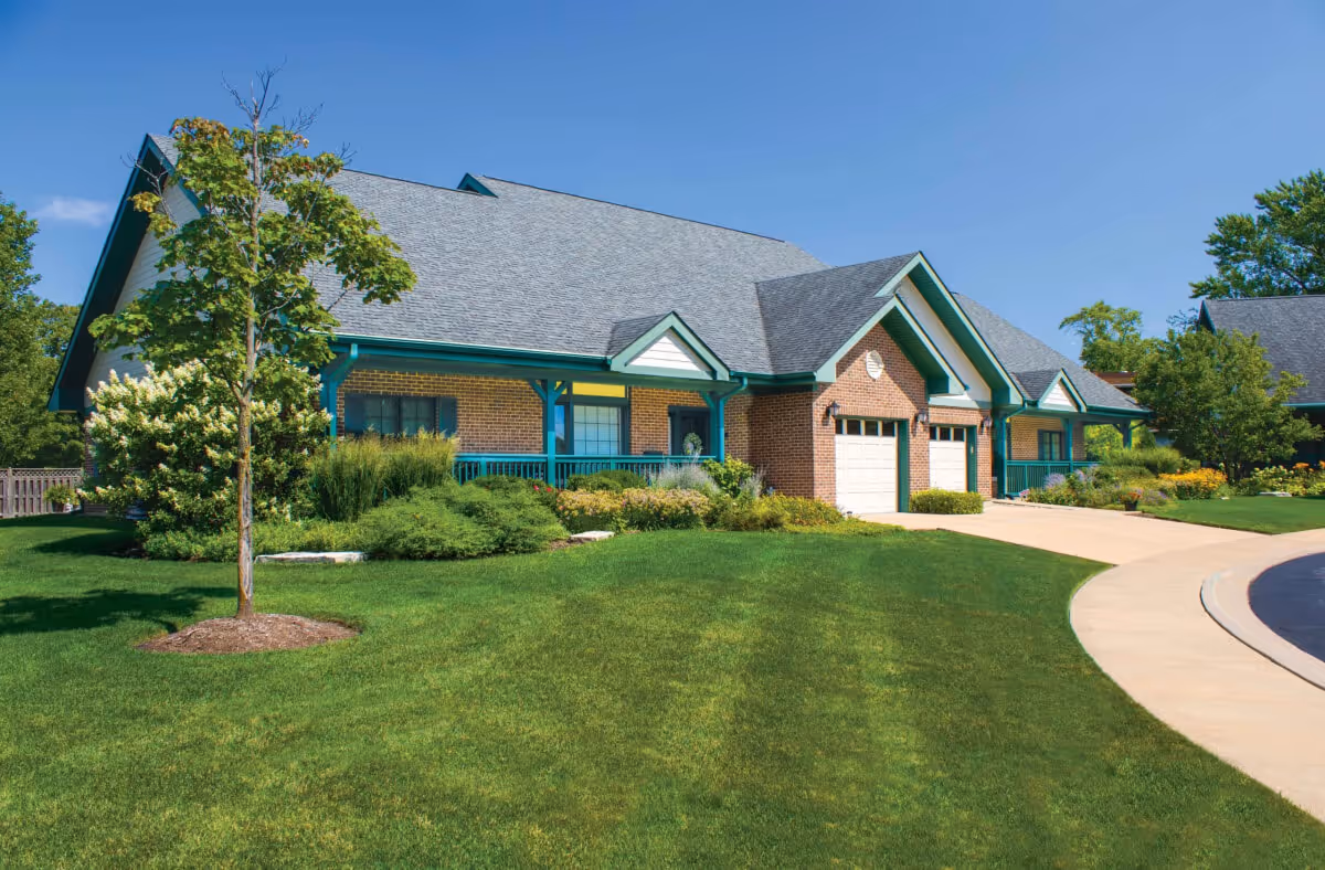 Brick single-story senior living building with green trim, attached garages, and a manicured lawn under a clear blue sky.