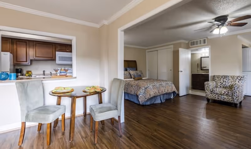 Interior view of a senior living facility showing a small dining area with a round wooden table set for two and two upholstered chairs. Adjacent to the dining area is a bedroom with a bed covered in patterned bedding, a closet with sliding doors, and a bathroom visible through an open door. There is also a patterned armchair and a ceiling fan with lights. The kitchen with wooden cabinets and white appliances is partially visible through a pass-through window.