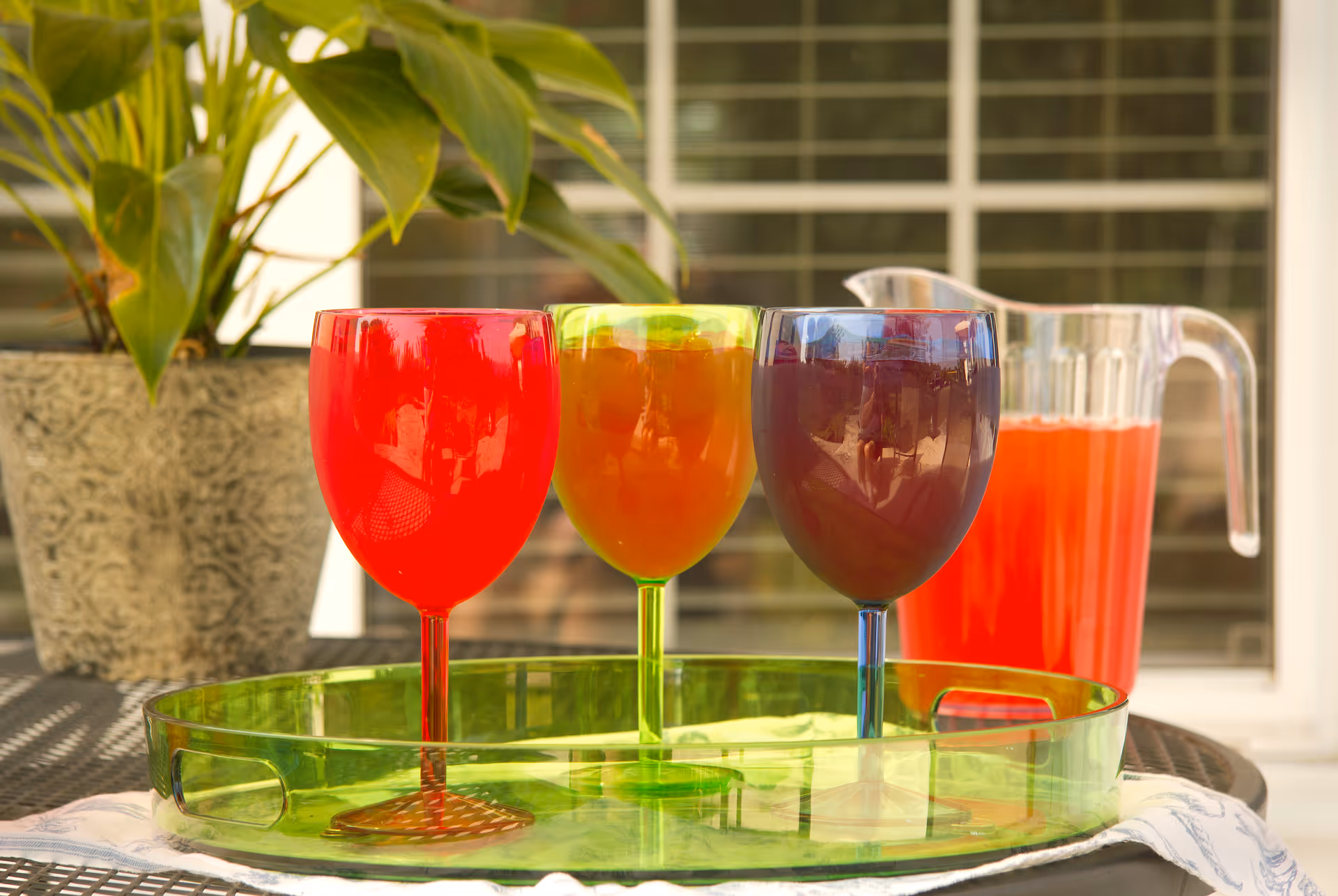 Three colorful translucent wine glasses filled with different beverages placed on a green tray on a table outdoors, with a pitcher of red drink and a potted plant in the background.