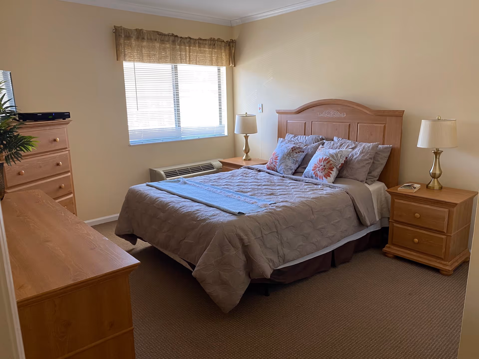 A neatly made bed with a wooden headboard in a bedroom. The bed has a gray quilted comforter, multiple pillows with floral patterns, and a folded blanket at the foot. On either side of the bed are wooden nightstands with matching lamps. There is a window with blinds and a valance above the bed, and wooden dressers are visible on the left side of the room. The walls are painted light beige and the floor is carpeted.