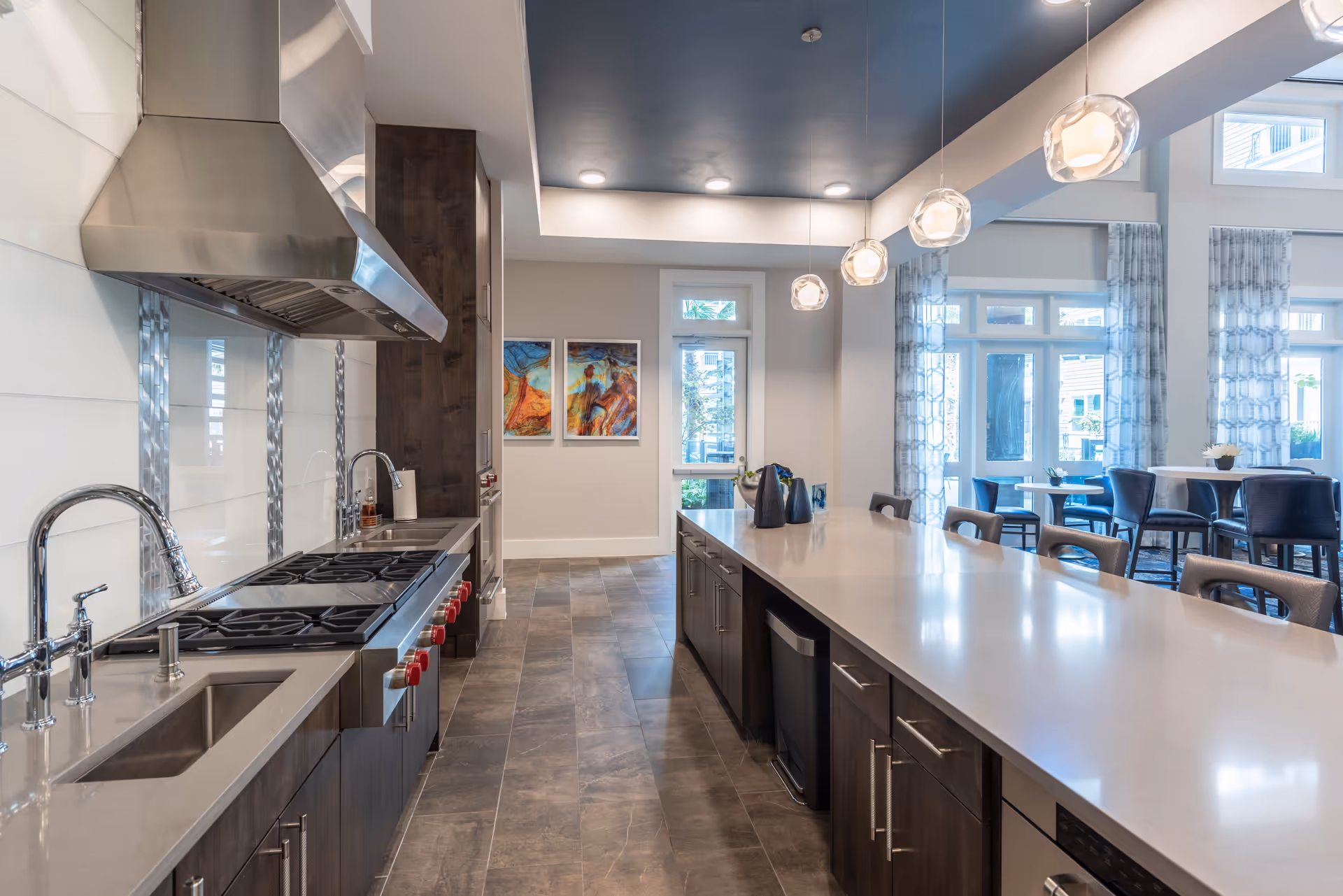 Modern communal kitchen with a long island, stovetop, pendant lights, and seating by large windows.